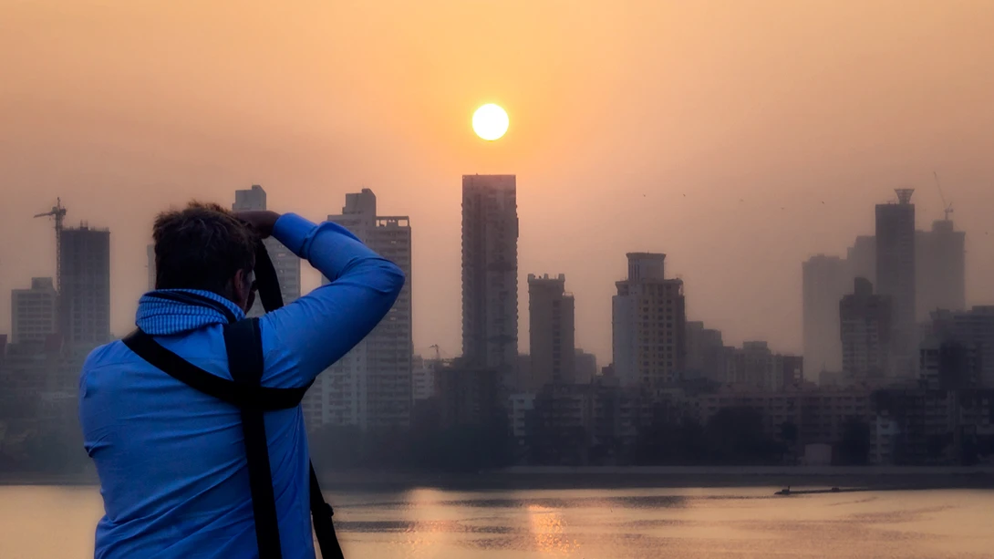 Photographer in blue jacket photographing sunrise over a city skyline reflected in water.