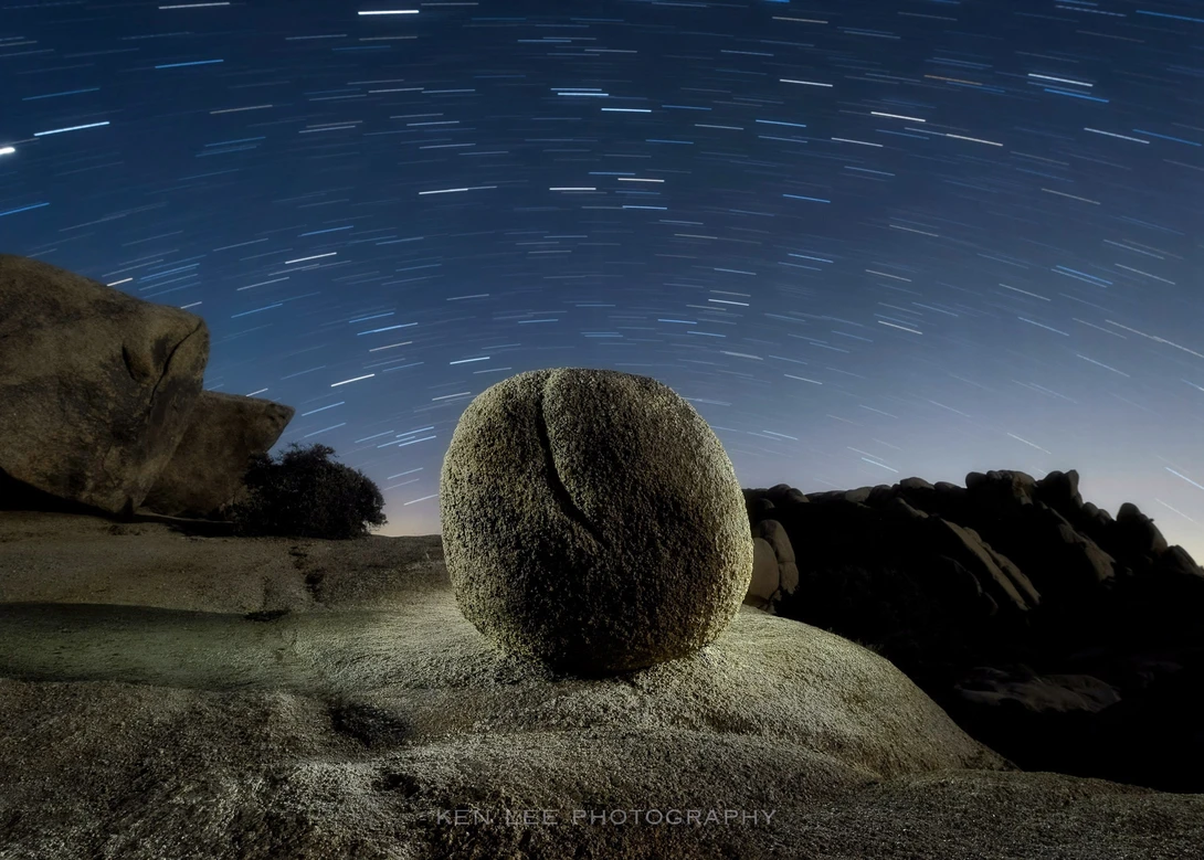 Large spherical boulder illuminated under star trails in a desert landscape at night.