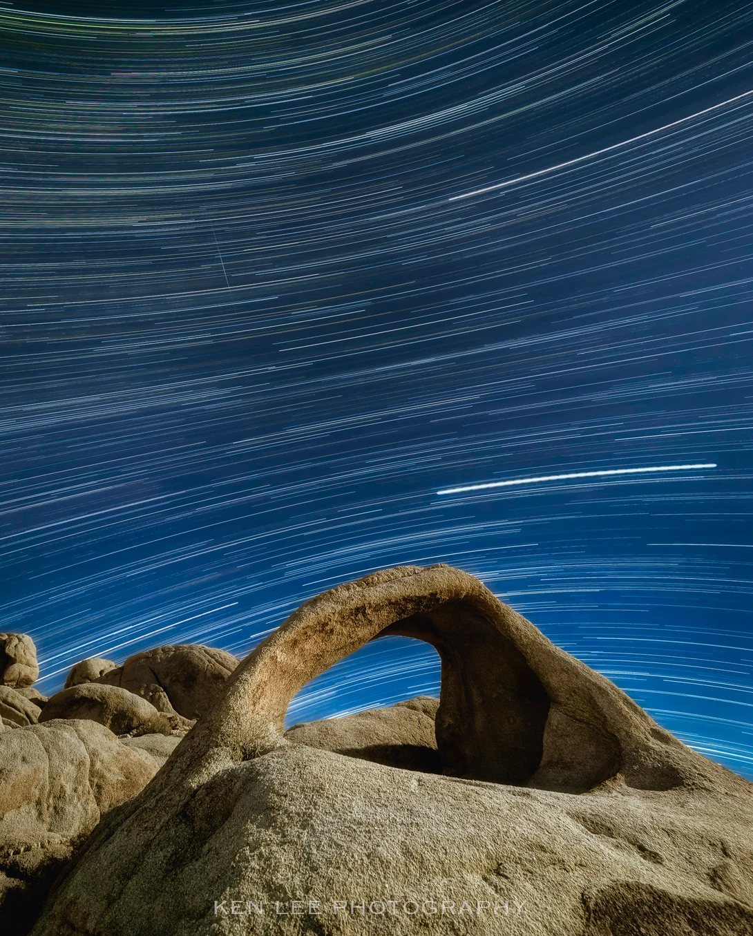 Large natural rock arch framing star trails streaking across a deep blue night sky.