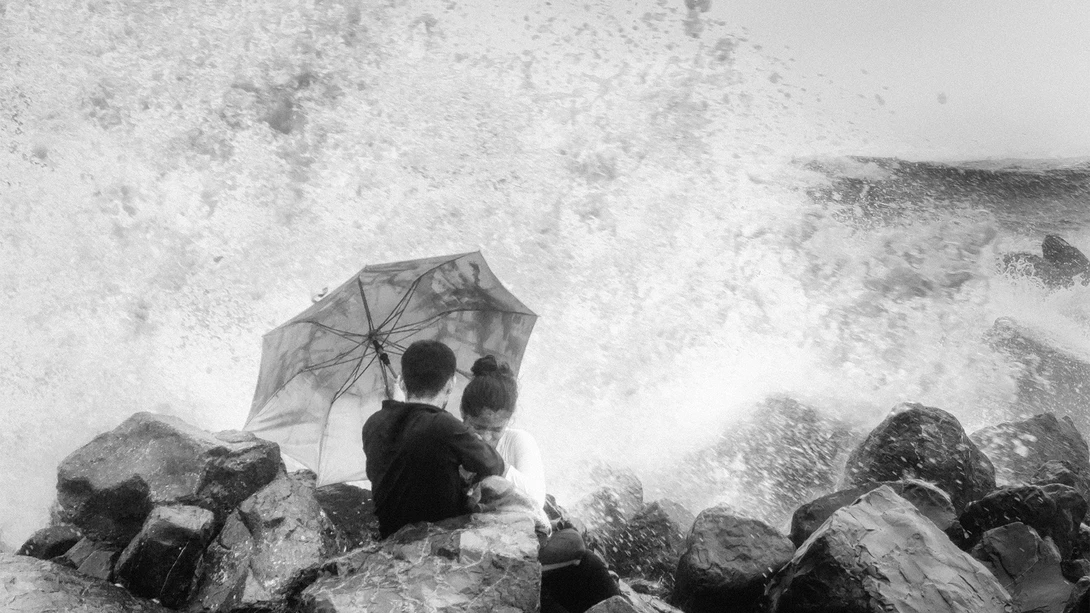 Person with umbrella standing on rocks facing large ocean waves in black and white.