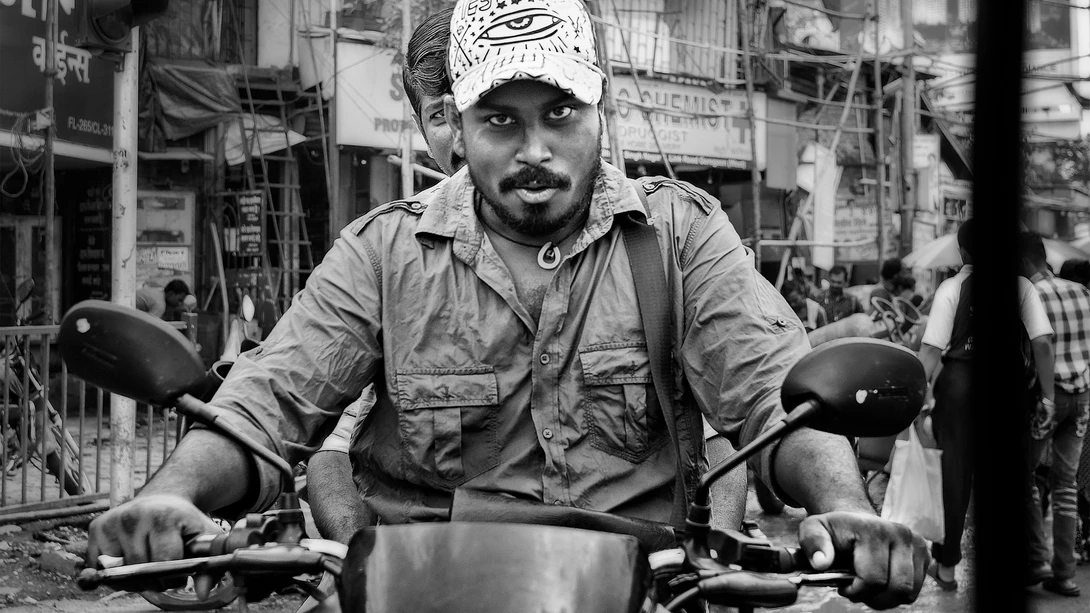 Man on motorcycle wearing cap and casual shirt, looking at camera in busy urban street.