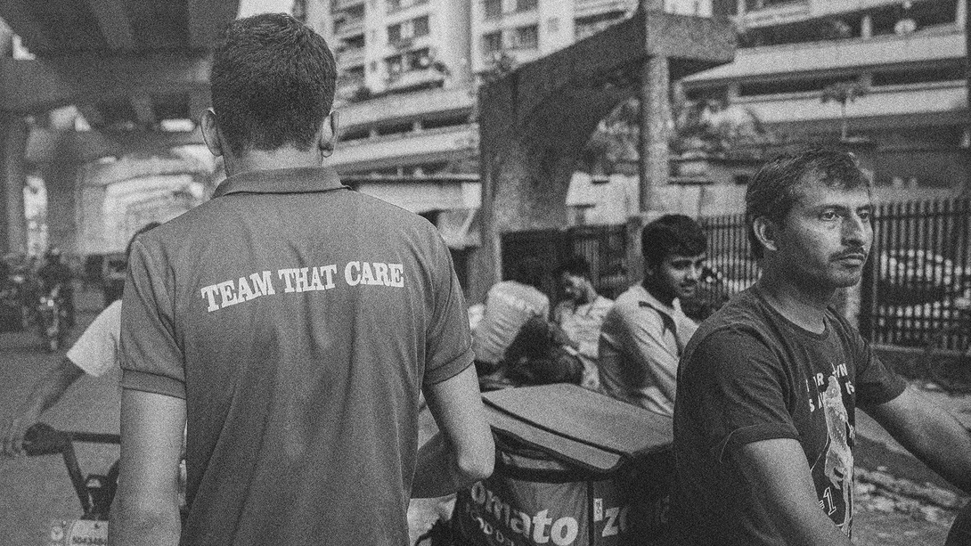 Man wearing Team That Care shirt stands in urban outdoor setting with other people visible in background.