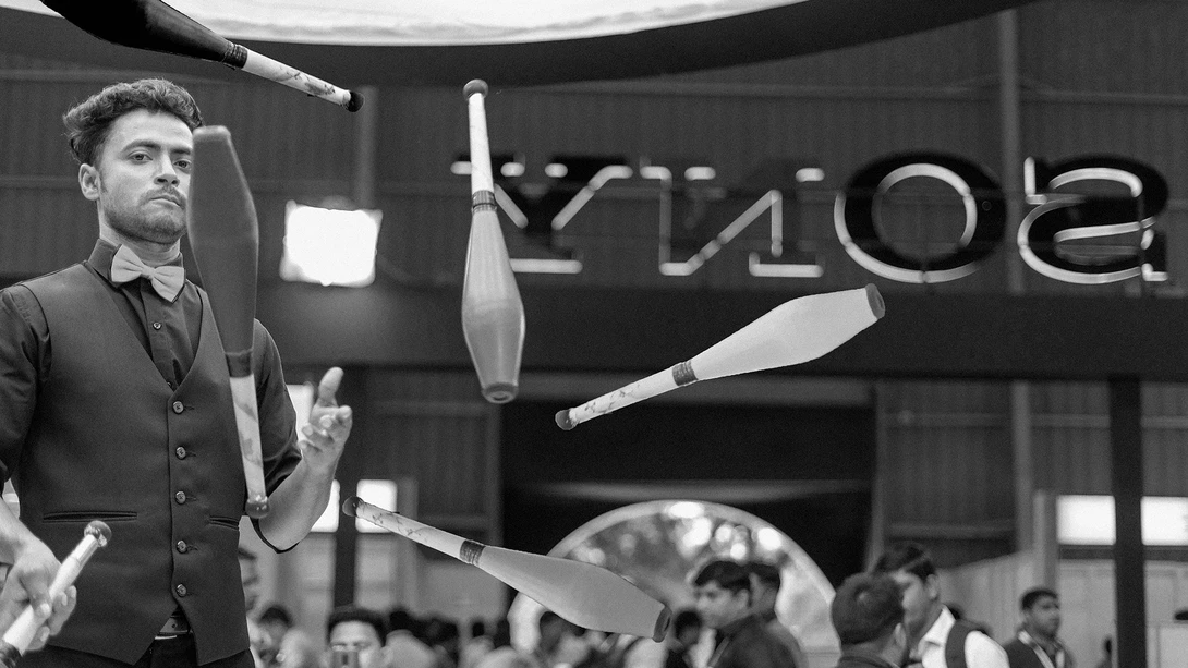 Juggler in dark vest performing with multiple clubs in mid-air during indoor performance.