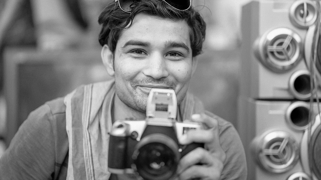 Photographer holding a vintage film camera to his face, smiling at the camera in black and white.