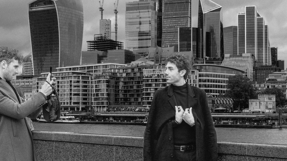 Man in dark jacket being photographed by another man holding a camera, with urban waterfront and skyscraper skyline behind them.