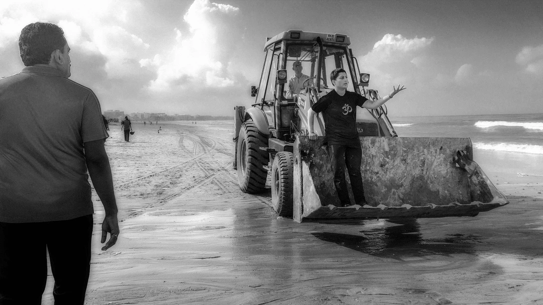 Person in dark shirt walking toward an industrial bulldozer on a wet beach.