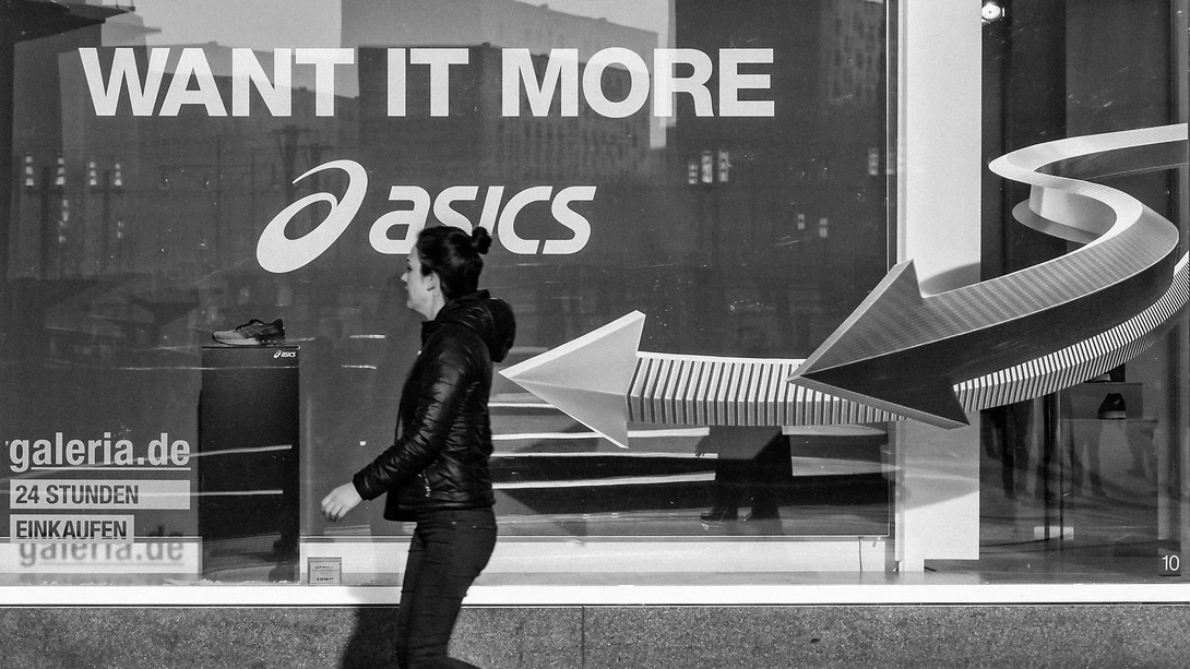 Person in black leather jacket walking past storefront window with Asics sportswear advertisement and geometric white design elements.