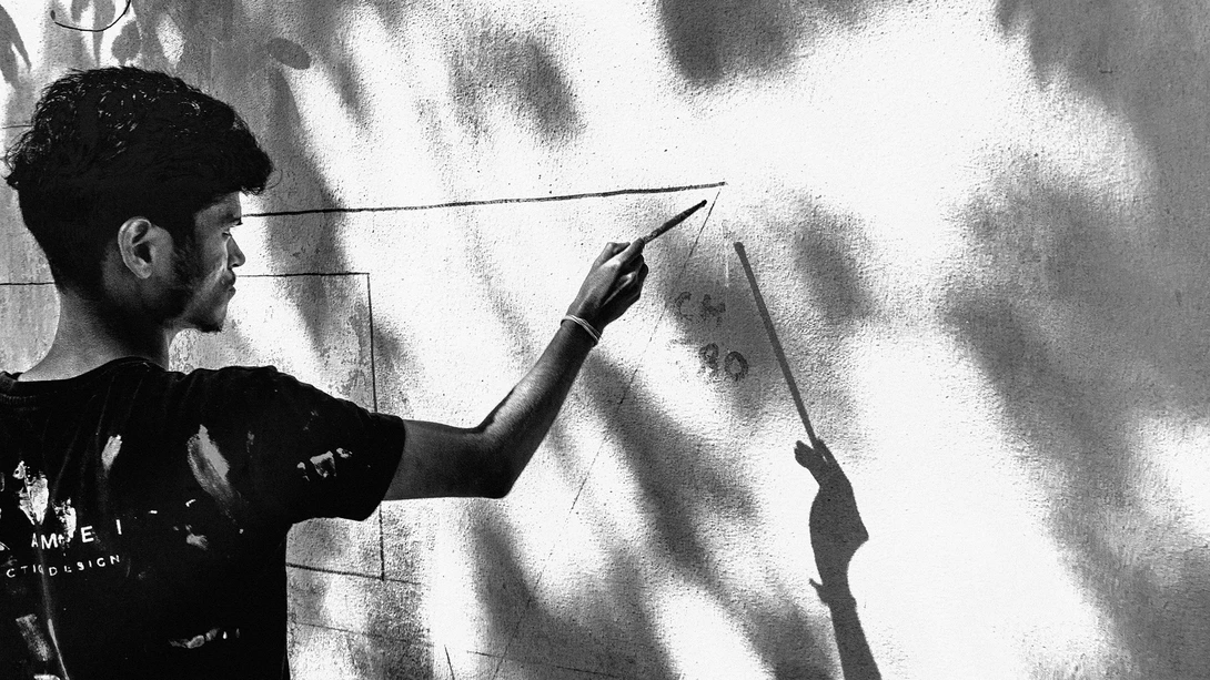 Artist drawing geometric lines on a white wall with a pen, casting dramatic shadows in natural light.