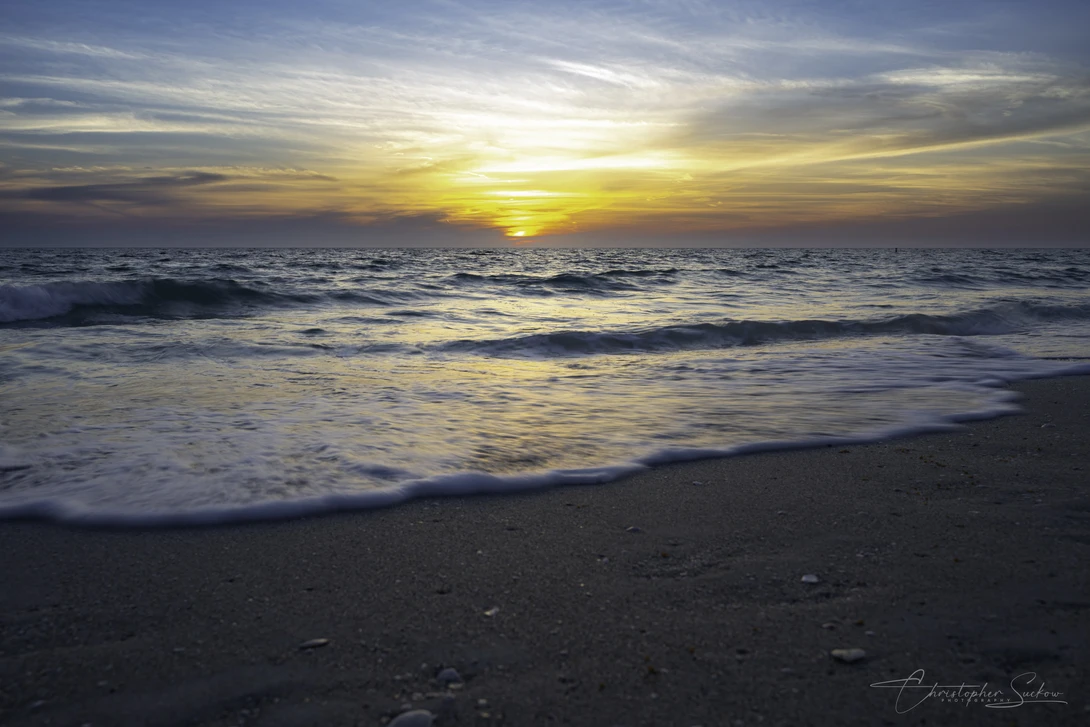 Golden sun setting over rolling ocean waves, with foamy surf washing across a wet sandy beach in the foreground.
